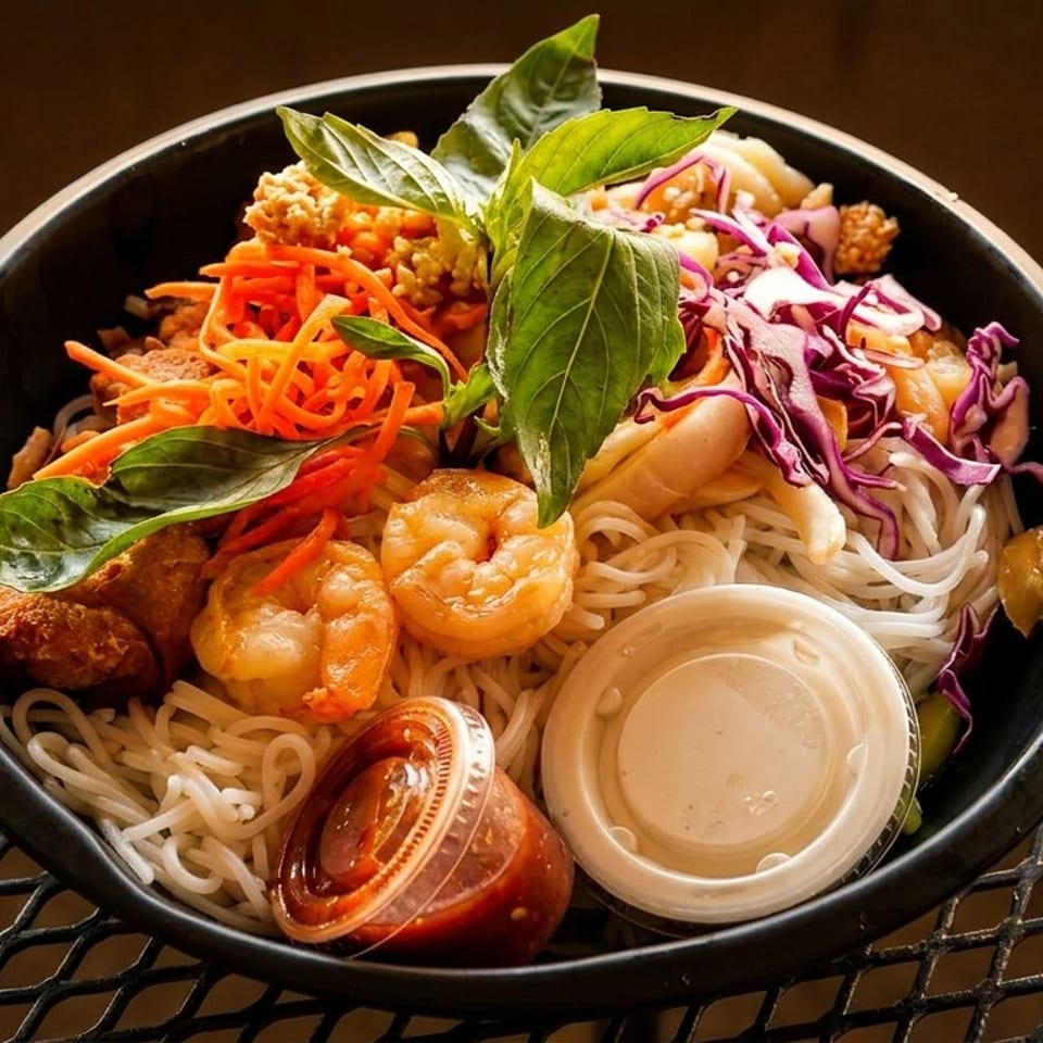 Colorful noodle bowl with shrimp, shredded carrots, purple cabbage, and fresh basil on a metal table.
