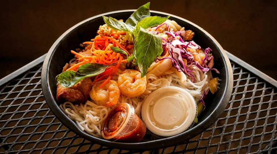A colorful noodle bowl with shrimp, shredded carrots, purple cabbage, and basil on a metal grate.