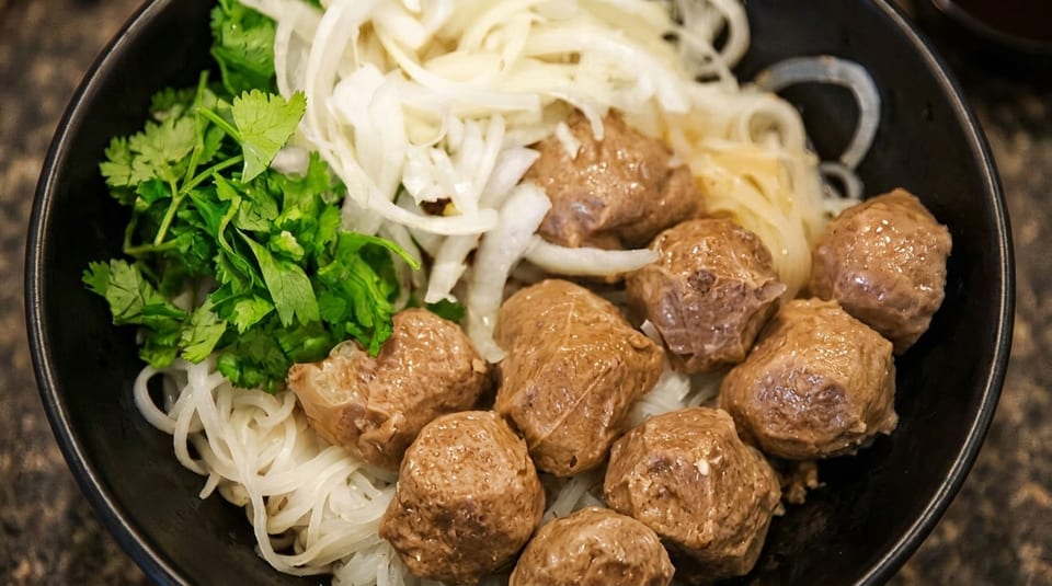 Close-up of a bowl of pho with meatballs, rice noodles, sliced onions, and fresh cilantro.