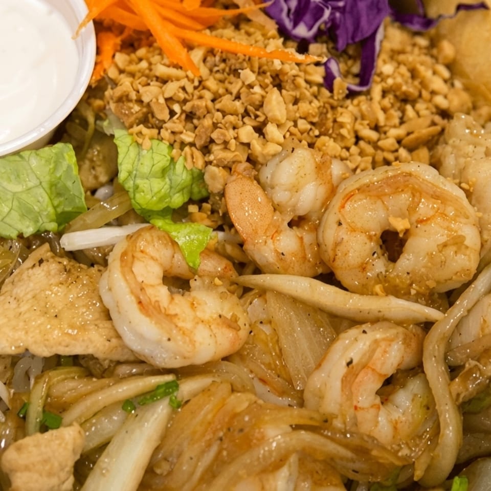 Close-up of a shrimp and chicken noodle dish with peanuts, shredded carrots, and a side of sauce.