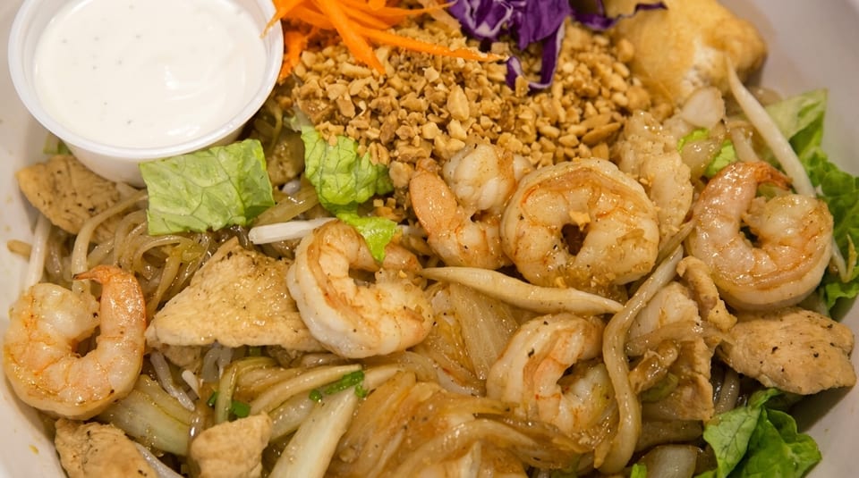 Close-up of a shrimp and chicken noodle dish with peanuts, shredded carrots, and a side of sauce.