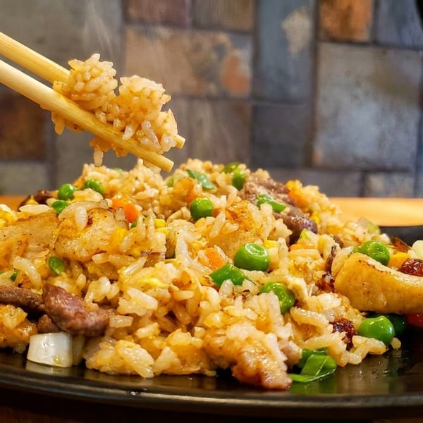 Chopsticks lifting steaming fried rice with peas, egg, and meat from a black plate.