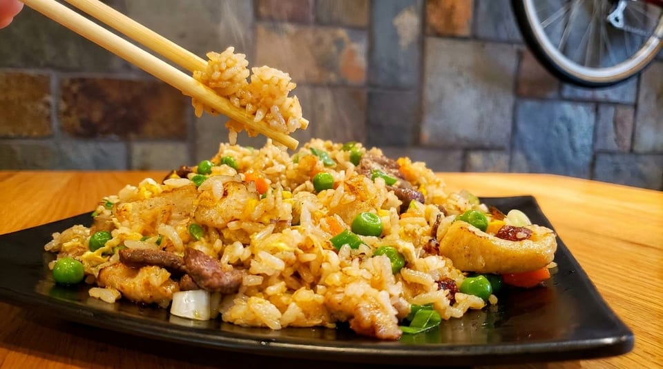 Chopsticks lifting steaming fried rice with peas, egg, and meat from a black plate.